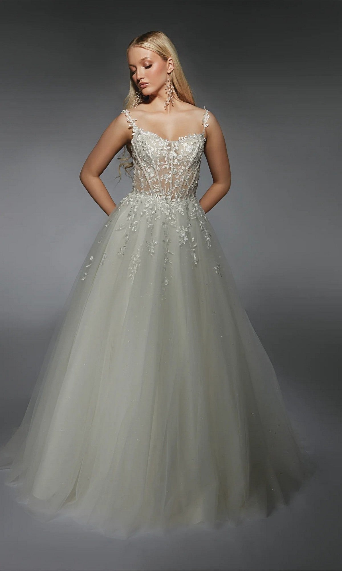 A woman models the Alyce 7130 long white dress in ivory/champagne, featuring a lace bodice and flowing tulle skirt, standing against a gray background.